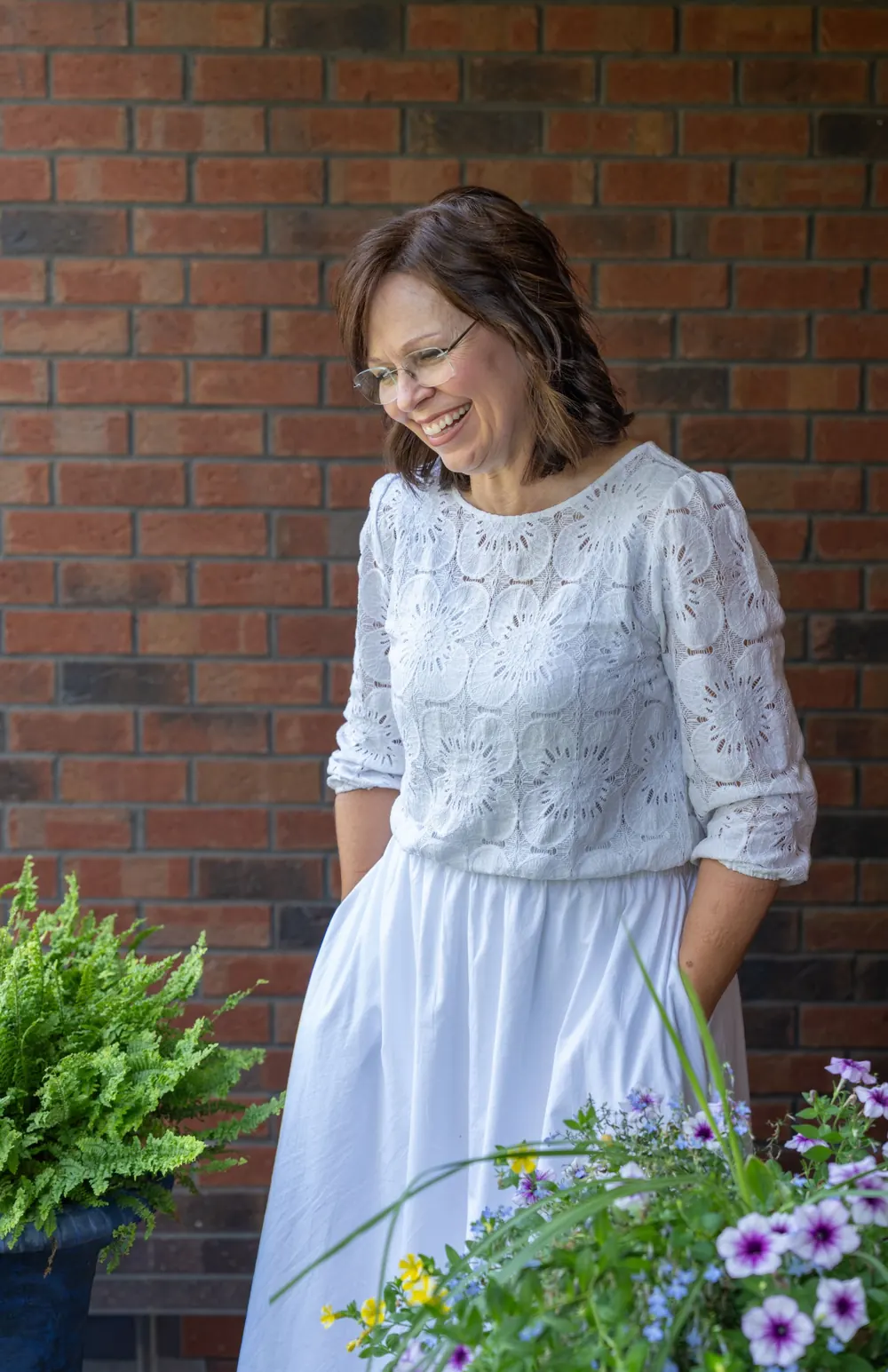 Jody Powers laughing, standing beside flowering planters in a white lace top and white skirt